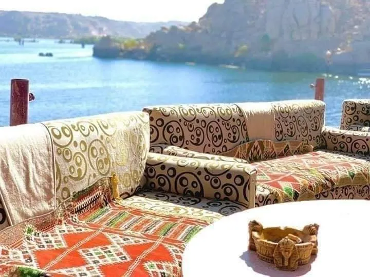 A vibrant seating area with patterned cushions overlooks the calm blue Nile River and desert cliffs in Aswan, Egypt.