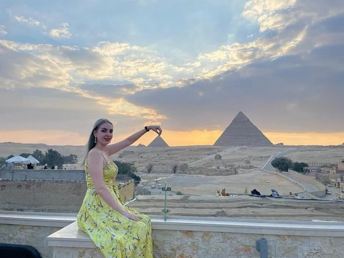 A woman in a yellow floral dress sitting on a balcony with a panoramic view of the Giza Pyramids at sunset.
