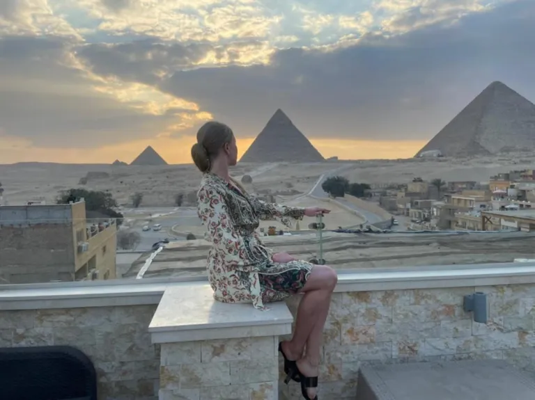 A woman sitting on a terrace looking at the Great Pyramids of Giza during a cloudy sunset in Cairo, Egypt.