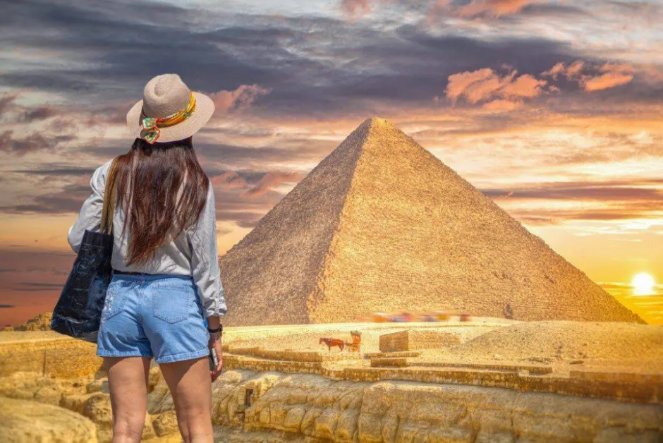 Back view of a woman wearing a hat looking at the Great Pyramid of Giza during a golden sunset in Egypt.