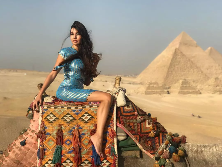 A woman in a blue dress sitting on a decorated camel with the Giza Pyramids in the background under a clear sky.