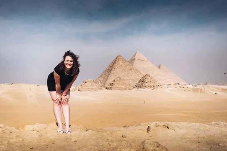 A smiling woman in a black dress posing in front of the three Great Pyramids of Giza on a sunny day in Egypt.