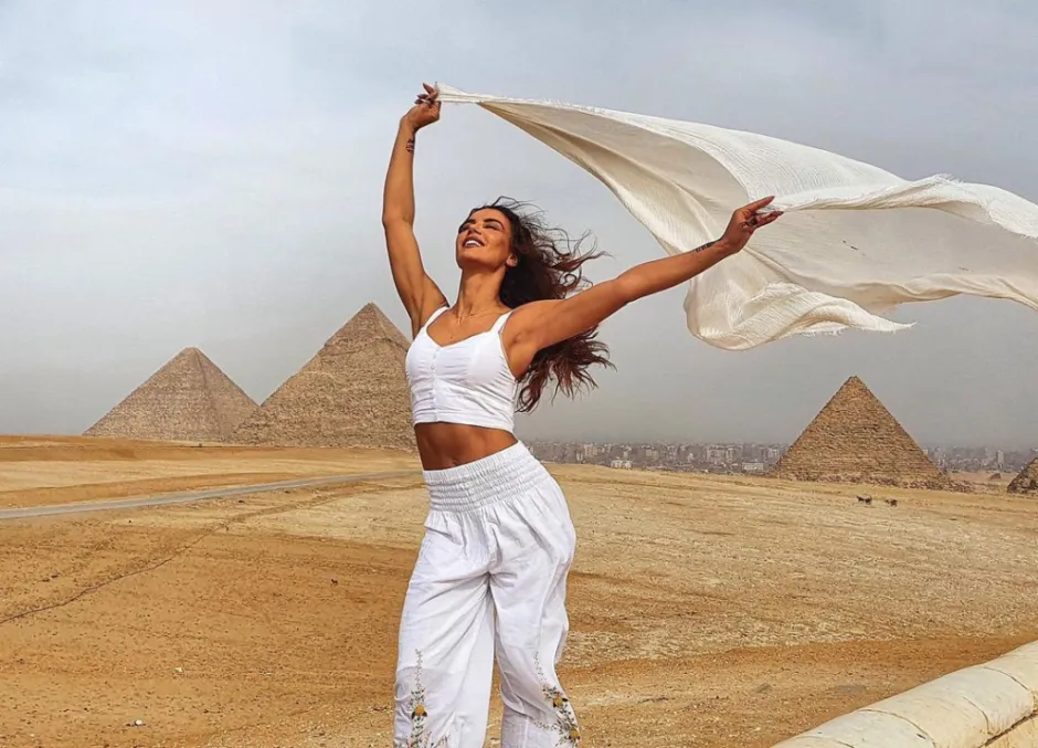 Smiling woman in white holding a flowing shawl with the Great Pyramids of Giza, Egypt, in the background under a cloudy sky.