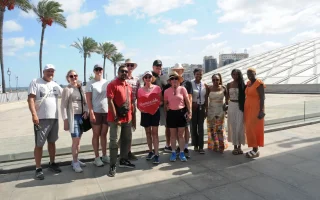 A diverse group of tourists posing with their guide in front of the modern Bibliotheca Alexandrina in Egypt. One woman holds a red heart-shaped sign, with palm trees and a sunny blue sky in the background.