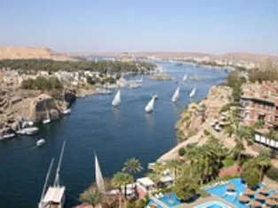 Traditional sailing feluccas on the Nile River in Aswan passing rocky islands, tropical greenery, and scenic desert landscapes.