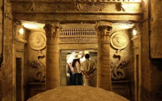 Tourists exploring the ancient Roman-Egyptian interior of the Catacombs of Kom El Shoqafa in Alexandria, featuring ornate pillars and carved wall reliefs of snakes.