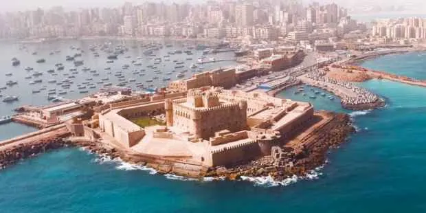 An aerial panoramic view of the 15th-century Citadel of Qaitbay fortress situated on the Mediterranean coast of Alexandria, Egypt, surrounded by turquoise waters and a harbor filled with small boats.