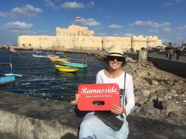 A happy tourist holding a Ramasside Tours sign in front of the Citadel of Qaitbay and colorful fishing boats in Alexandria, Egypt.