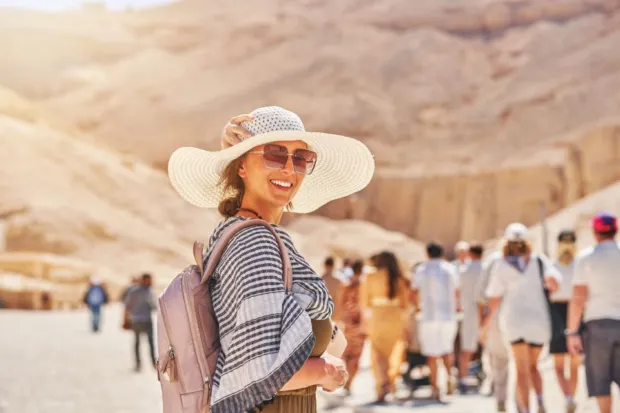A smiling woman in a sun hat and sunglasses exploring the desert mountains of Luxor, Egypt, with tourists behind.