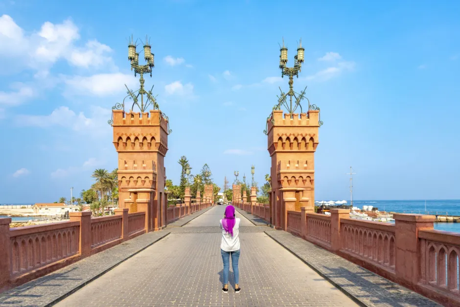 A tourist standing on the historic Montaza Palace bridge in Alexandria, Egypt, overlooking the Mediterranean Sea and lush royal gardens.