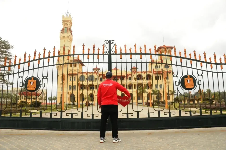 A professional tour guide standing before the ornate gates of the historic Montaza Palace in Alexandria, Egypt, explaining the history of its distinctive Florentine-inspired tower and royal gardens.