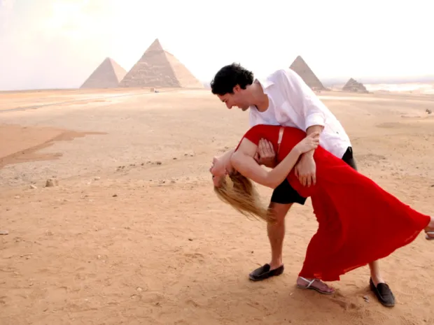 A man dipping a woman in a red dress during a romantic dance in the desert with the Giza Pyramids in the background.
