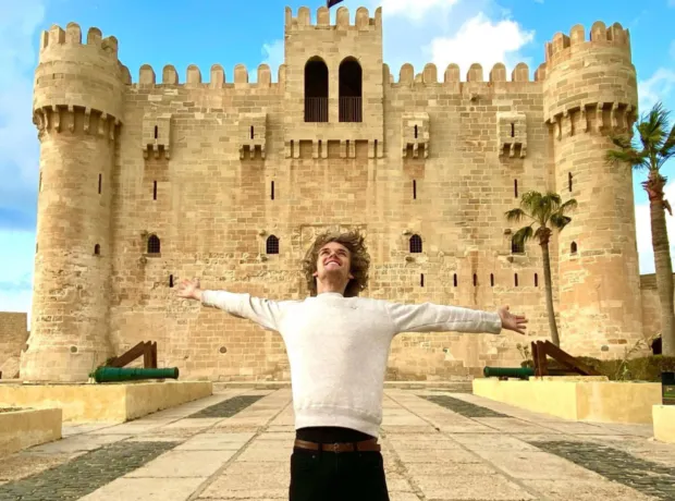 A young male tourist standing with arms wide open in front of the historic Citadel of Qaitbay in Alexandria, Egypt, featuring the ancient stone walls and defensive towers.