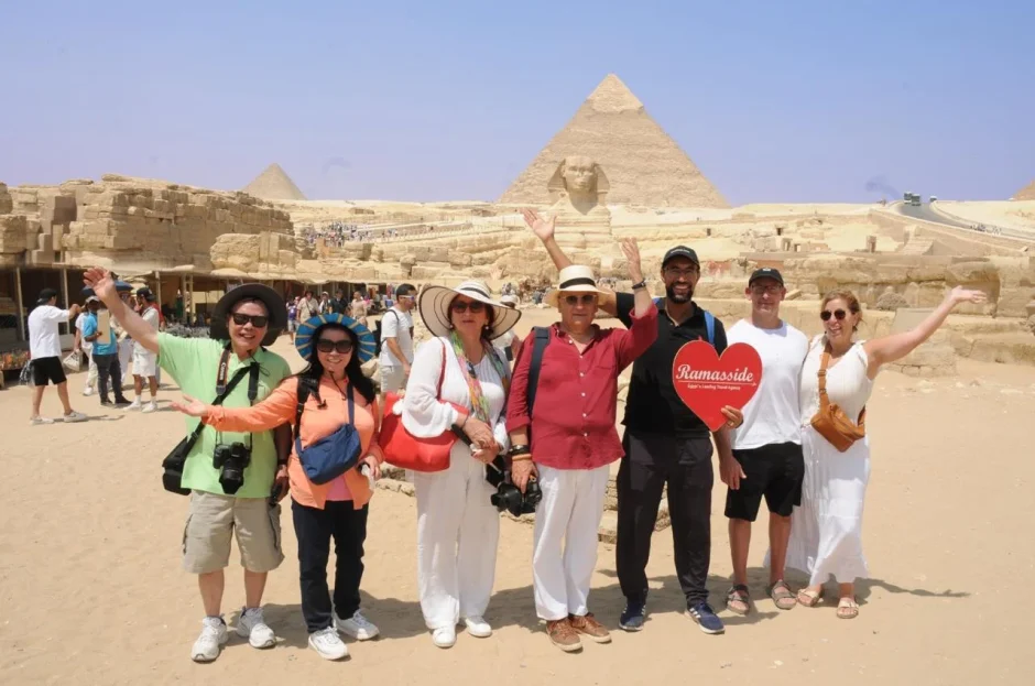 A group of happy tourists posing in front of the Great Sphinx and Giza Pyramids in Egypt under a clear blue sky.