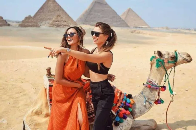 Two happy women enjoying a camel ride vacation at the Giza Pyramids, pointing at the historic landmarks in the Egyptian desert.
